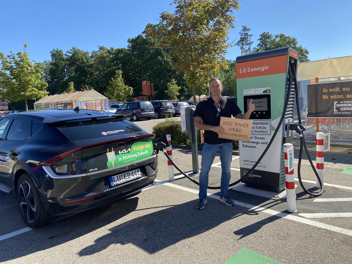 Elektroauto von Pfalzwerke lädt an einer öffentlichen Ladesäule; daneben steht eine Person mit Schild ‚Volle Power voraus‘ auf einem Parkplatz im Freien.